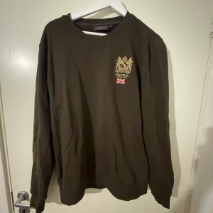Brun sweatshirt från Morris, storlek L - Säljer en brun sweatshirt från Morris i storlek L. Tröjan har rund halsringning, lång ärm och ett broderat emblem med brittisk flagga på bröstet. Perfekt för dig som gillar klassisk och stilren design.