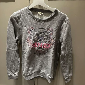 Grå Kenzo sweatshirt med tiger - Säljer en grå sweatshirt från Kenzo med ikoniskt broderat tigerhuvud och rosa Kenzo-logga på bröstet. Tröjan har rund hals, långa ärmar och är gjord i mjuk bomull. Perfekt för dig som vill sticka ut med en streetig vibe.