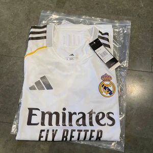 Real Madrid hemmatröja 25/26 L - Snygg vit Real Madrid hemmatröja från Adidas med svarta detaljer och gula inslag längs sidorna. Klassisk klubbmärke på bröstet, Adidas-logga och sponsortryck framtill. Tröjan är kortärmad och tillverkad i lätt, ventilerande material (players version). Inget namn på ryggen, nyskick