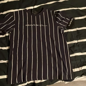 Svart randig t-shirt Kings Will Dream - Snygg svart t-shirt från Kings Will Dream med vita vertikala ränder och logga broderad på bröstet och ärmslutet. Klassisk rund hals och korta ärmar. Perfekt för dig som gillar streetwear och vill sticka ut med stilren design.