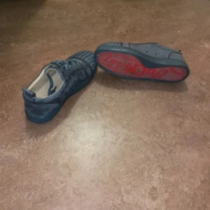 Christian Louboutin sneakers med nitar - Säljer ett par mörkgrå sneakers från Christian Louboutin i mocka med ikonisk röd sula och coola nitar över tån. Skorna har snörning och en rund tå, perfekt för dig som vill sticka ut med din stil.