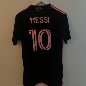 Inter Miami Messi fotbollströja - Snygg svart Inter Miami fotbollströja från Adidas med rosa detaljer och Messi #10 på ryggen. Tröjan har klubbmärke, sponsortryck och klassiska Adidas-ränder på axlarna. Tillverkad i lätt och ventilerande material, perfekt för match eller träning. (ÄKTA) köpt på adidas hemsida för 1099kr.