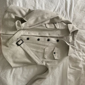  kappa från NLY Trend - Stilren beige kappa från NLY Trend i storlek 32. Jackan har klassisk krage, svarta knappar framtill, två bröstfickor med lock och ett matchande bälte i midjan. Perfekt för dig som gillar en clean och tidlös look.