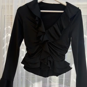 Svart omlottopp med volang - Snygg svart omlottopp med långa ärmar och volangdetaljer längs framstycket. Toppen har en figurnära passform och är tillverkad i ett mjukt, stretchigt material som känns bekvämt mot huden. Perfekt för dig som gillar en stilren look med extra detaljer.