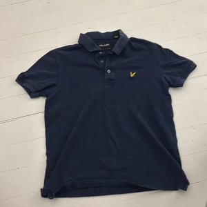 Marinblå Lyle & Scott pikétröja - Snygg marinblå piké från Lyle & Scott med klassisk krage och tre knappar framtill. T-shirten har korta ärmar och är gjord i mjuk bomull. På bröstet sitter den ikoniska gula fågeln broderad. Perfekt för en chill och stilren look.