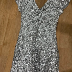 Glittrig silverklänning från Oneness - Säljer en kort, glittrig klänning från Oneness i silver med paljetter över hela plagget. Klänningen har v-ringning och korta ärmar, perfekt för dig som vill sticka ut. Materialet är syntet och den har en figurnära passform.