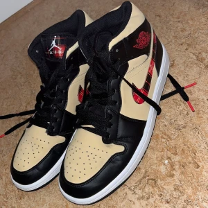 Nike Air Jordan 1 Mid Beige/Black Rutig - Nike Air Jordan 1 Mid i beige och svart skinn med röd-rutiga detaljer på swoosh och plös. Klassisk siluett med svart snörning, vit mellansula och svart yttersula.  med en unik färgkombo och rutigt mönster helt nya NYPRIS LIGGER PÅ 1900kr
