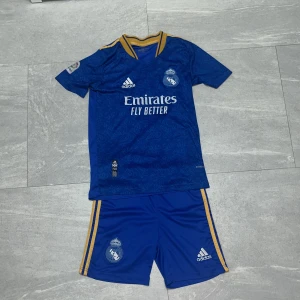 Real Madrid set  - Säljer en blå Real Madrid matchtröja med Benzema och nummer 9 på ryggen. Tröjan har korta ärmar, gul krage och gula ränder på axlarna. Materialet är lätt och andas, perfekt för fotboll. Setet inkluderar matchande blå shorts med gula ränder och klubbmärke. Shorts för 75kr, båda för 200kr