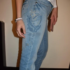 Ljusblå jeans med broderi och bootcut - Säljer ett par ljusblå jeans med snyggt broderi på låret och bakfickan. Jeansen har bootcut-modell. Innebredslängd 78cm och tvärs över 34cm.