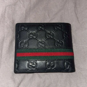 Svart Gucci plånbok med GG-mönster - Snygg svart plånbok från Gucci med präglat GG-mönster och klassisk grön-röd rand på framsidan. Plånboken är i skinn och har flera fack för kort och sedlar. Perfekt för dig som vill ha en lyxig och stilren accessoar.