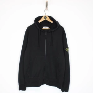 Svart zip hoodie från Stone Island - Svart hoodiejacka från Stone Island med dragkedja framtill och klassisk patch på vänster ärm. Jackan har huva med dragsko, två fickor och ribbade muddar. Perfekt för dig som gillar streetwear och vill ha en stilren look.