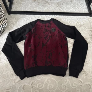 Vinröd och svart tröja från Philipp Plein - Cool vinröd och svart tröja från Philipp Plein med unikt mönster framtill och svart logomärke på bröstet. Tröjan har svarta ärmar och muddar, rund halsringning och en loose passform. Perfekt för dig som vill sticka ut med en streetstyle-look.