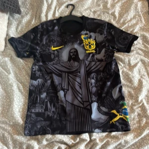 Svart Brasil Nike fotbollströja - Unik svart Brasilien fotbollströja från Nike med detaljerat grafiskt tryck av Kristusstatyn och brasilianska motiv på framsidan. Gula loggor och emblem, samt inslag av blått och grönt på ärmarna. Tillverkad i lätt och ventilerande material, perfekt för match eller träning.