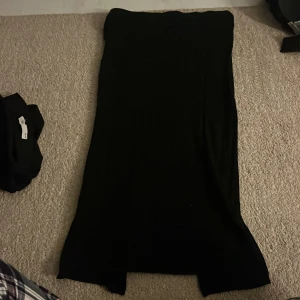 Svart långklänning från The Black Label - Säljer en svart axelbandslös långklänning från The Black Label Collection i storlek M. Kjolenhar en stilren och enkel design med slits nertill för extra rörelsefrihet. Perfekt för dig som gillar minimalistisk och elegant stil.