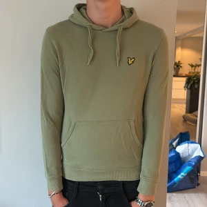 Olivgrön hoodie från Lyle & Scott - Snygg olivgrön hoodie från Lyle & Scott med klassisk gul logga på bröstet. Tröjan har huva med snörning, känguruficka och ribbade muddar. Perfekt för en chill och stilren look.