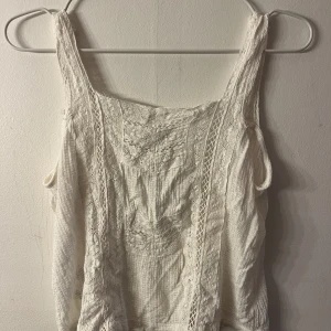 Vit spetsig linnetopp med broderi - Superfin vit linnetopp med breda axelband och detaljerad spets längs hela toppen. Broderade mönster framtill och inslag av virkad spets ger en romantisk känsla. Perfekt till sommarens alla outfits!
