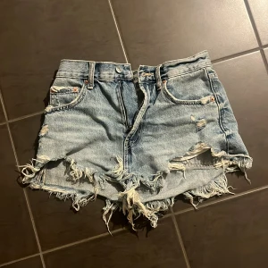 Blå jeansshorts med fransar från Zara - Säljer ett par blå jeansshorts från Zara i storlek 34. De är högmidjade med råa, fransiga kanter och slitna detaljer för en cool look. Klassisk femficksmodell med dragkedja och knapp. Perfekta till sommaren!