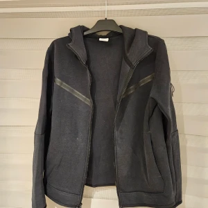 Svart Nike hoodiejacka med dragkedja M - Nike hoodiejacka i svart med dragkedja hela vägen, huva och snygga diagonala detaljer på bröstet. Jackan har fickor på sidorna och en extra ficka med dragkedja på ärmen. Perfekt för chill dagar eller när du vill ha en sportig look.