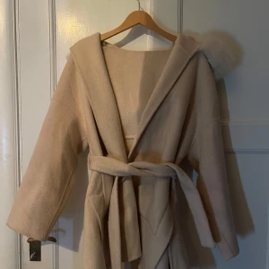 Beige kappa med huva och päls - Säljer en beige kappa med stor huva och avtagbar päls runt kanten. Jackan har knytskärp i midjan, stora fickor framtill och är långärmad. Perfekt för dig som vill ha en mysig och stilren look under kyliga dagar. Stolek S från luxeswe