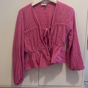 Rosa blus med knyt från ASOS DESIGN - Supersöt rosa blus från ASOS DESIGN med långa ärmar, knyt framtill och volang nertill. Blusen har en lätt rynkad struktur, knappar framtill och v-ringad hals. Perfekt för dig som gillar färg och en avslappnad men trendig look.
