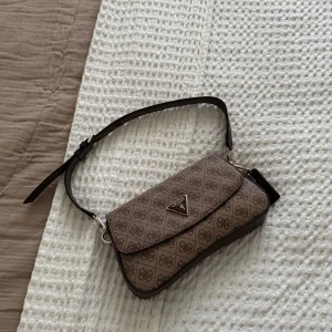 Beige axelväska från Guess - Säljer en snygg beige axelväska från Guess med klassiskt logomönster och justerbar rem. Väskan har silverfärgade detaljer, dragkedja på insidan och dekorativa tags med 'Guess' och '1981'. Perfekt storlek för det viktigaste och riktigt trendig look.