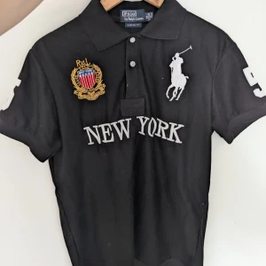 Svart Chief keef Pike med New York tryck - Det svart chief keef piké från Ralph Lauren med ljusblått New York-tryck och nummer 5 på ryggen. Tröjan har korta ärmar och en klassisk krage. Säljer pga att man bytt stil