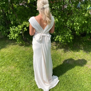 Vit långklänning med rosett i satin - Supersnygg vit långklänning i glansigt satinliknande material. Djup v-ringning fram och öppen rygg med en stor rosett bak som ger en extra elegant touch. Klänningen har en figurnära passform och faller snyggt längs kroppen. Perfekt för studentbal!💗