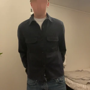 Mörkblå overshirt med fickor - Snygg mörkblå overshirt med klassisk krage och två stora bröstfickor med lock. Skjortan har knäppning framtill och är långärmad. Perfekt att bära över en t-shirt för en avslappnad men stilren look.