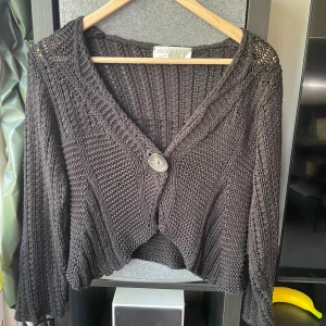 Svart stickad kofta från Joachim Bosse - Säljer en svart stickad kofta från Joachim Bosse med grovstickad struktur och en stor, unik knapp framtill. Koftan har v-ringad hals, långärmade och lite kortare passform som ger en chill vibe. Perfekt att slänga över en t-shirt när det blir lite kyligt.