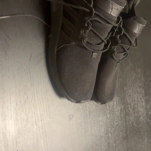 Svarta Adidas sneakers med mesh - Snygga helsvarta Adidas sneakers med ovandel i mesh och klassiska tre ränder på sidan. Skorna har snörning och en sportig vibe, perfekt för dig som gillar stilrena och bekväma sneakers. Adidas-logga på plösen och detaljer i syntet.