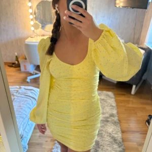 Gul blommig kortklänning med puffärm  - Supersöt gul kortklänning med små blommor, puffiga långärmar och snygg snörning i ryggen från Nelly. Klänningen har rynkade detaljer och en feminin, figurnära passform. Perfekt för dig som vill sticka ut med färg och volym på ärmarna. Materialet känns lätt och somrigt.