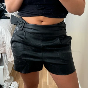 Svarta högmidjade skinnshorts med bälte - Snygga svarta shorts i skinnimitation med hög midja och ett brett bälte med stor spänne. Shortsen har sidfickor och en diskret dragkedja i sidan men som är sönder men ingen som läggs märke till. Passformen är loose och de har en cool, edgy vibe som passar perfekt till en croppad topp.