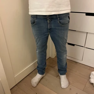 Blå Replay jeans  - Säljer ett par blå jeans från Replay med snygg tvätt och klassiska fem fickor. Jeansen har tapered passform och normal midja, med diskreta slitningar för en avslappnad look. Har en slitning vid rumpan men det är inget man märker.