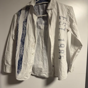 Vit skjorta med blåa ränder från Lindex - Säljer en vit långärmad skjorta från Lindex med coola blåa ränder och stjärnor på ryggen. Skjortan har trycket 'EST 1985' på ena ärmen och knäppning framtill. Perfekt för dig som vill sticka ut lite extra!