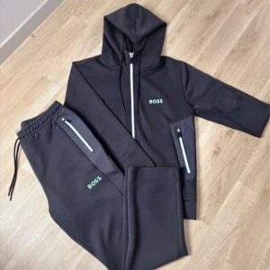 Mörkgrå BOSS Tracksuit - Mörkgrå hoodie från BOSS med hel dragkedja framtill, huva med snörning och limegröna detaljer vid fickor och dragkedja. Tröjan har lång ärm och en lime grön BOSS-logga på bröstet. Tillverkad i mjuk polyester, perfekt för en chill och sportig stil. Samma med byxorna. Helt Nya.