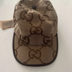 Beige keps från ggi med GG-mönster - Snygg beige keps från ggmed klassiskt brunt GG-mönster över hela kepsen. Kepsen har böjd skärm med mörkbrun kant och är tillverkad i ett slitstarkt tygmaterial. Perfekt för dig som vill sticka ut med en lyxig accessoar.