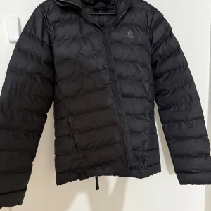 Svart pufferjacka från Adidas - Svart pufferjacka från Adidas med klassisk quiltad design och dragkedja framtill. Jackan har lång ärm, diskret logga på bröstet och reflekterande detaljer på huvan. Perfekt för kalla dagar och enkel att matcha med allt.