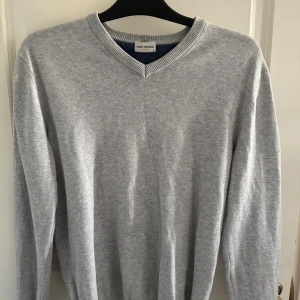 John Henric tröja  - Säljer en stilren ljusgrå stickad tröja från John Henric med v-ringad hals. Tröjan har lång ärm och är tillverkad i mjukt material som passar perfekt till lager-på-lager. Enkel och clean look som funkar till många outfits.