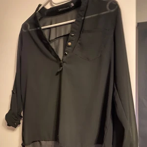 Svart henleyblus från Zanzea - Svart henleyblus från Zanzea med lång ärm och knappar framtill. Skjortan har en bröstficka och är tillverkad i ett lätt, flowy material som ger en snygg och avslappnad look. Perfekt att styla med jeans eller kjol för en clean vibe.