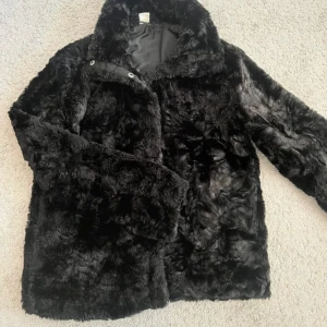 Svart pälsjacka med krage - Mysig svart pälsjacka med knäppning framtill. Jackan har en fluffig och lyxig känsla, perfekt för kalla dagar. Passformen är normal och jackan är helsvart utan mönster. Snygg att styla till både jeans och klänning och perfekt nu till hösten, den är även lite vindtät💕