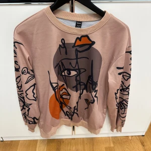 Beige sweatshirt med grafiskt tryck  - Cool beige sweatshirt från SHEIN med unikt grafiskt ansiktstryck i svart, grått och orange på både fram- och baksida. Tröjan har rund hals, långa ärmar och relaxed passform. 