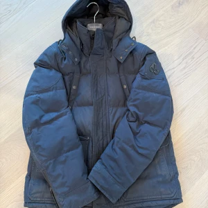 Mörkblå dunjacka Moose Knuckles - Snygg mörkblå dunjacka från Moose Knuckles med huva och ikonisk logga på ärmen. Jackan har flera fickor, dragkedja och knappar framtill. Perfekt för kalla dagar och riktigt street i stilen. Jackan är väll använd, har ägt den i några år nu 