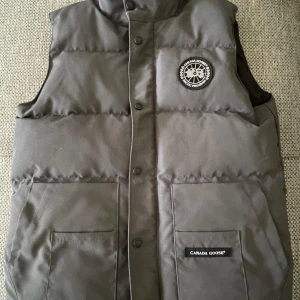 Grå dunväst från Canada Goose - Säljer en grå dunväst från Canada Goose med svart och vit logga på bröstet. Västen är i nyskick. Kan passa även storlek M