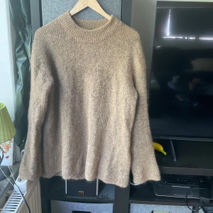 Beige stickad tröja By Malene Birger - Mysig beige stickad tröja från By Malene Birger med rund halsringning och lång ärm. Tröjan har en loose fit och är tillverkad i ett ulligt material som känns både lyxigt och varmt. Perfekt för lager-på-lager under kyliga dagar.