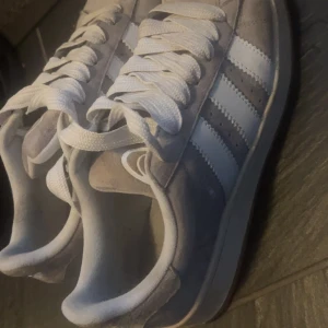 Grå Adidas campus  sneakers mocka - Snygga grå Adidas Gazelle sneakers i mocka med klassiska vita ränder på sidorna och vit sula. Skorna har rund tå, platt sula och vita snören. Perfekta för dig som gillar en clean och tidlös streetstil.