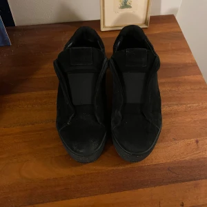 Svarta sneakers i mocka - Stilrena svarta sneakers i mocka med platt sula och rund tå. Skorna har en minimalistisk design utan synlig snörning och breda elastiska band över vristen för en clean look. Perfekta för dig som gillar enkel och modern stil.