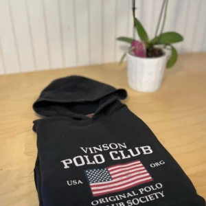 Svart hoodie från Vinson Polo Club - Svart hoodie från Vinson Polo Club med tryck framtill och amerikansk flagga. Klassisk huva och långa ärmar, ribbade muddar och mjukt material. Perfekt för dig som gillar sportig och avslappnad stil.