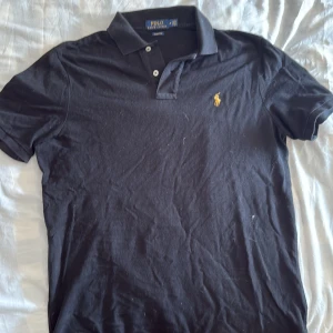 Svart pikétröja från Polo Ralph Lauren - Klassisk svart pikétröja från Polo Ralph Lauren med korta ärmar och gul broderad logga på bröstet. Tröjan har krage, två knappar vid halsen och är tillverkad i mjuk bomull. Perfekt för en stilren och avslappnad look.