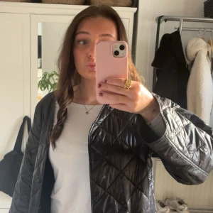 Svart quiltad jacka i loose fit - Säljer en svart quiltad jacka med loose passform. Jackan har ett stilrent rutmönster och är lätt vadderad, perfekt för lager-på-lager. Den har lång ärm och stängs med knappar framtill. Enkel att styla till både jeans och kjol.