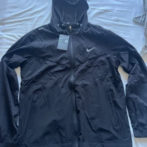 Nike vindjacka - Svart vindjacka från Nike med huva och reflekterande detaljer. Jackan har hel dragkedja framtill, två sidofickor och justerbar dragsko i huvan. Tillverkad i lätt polyester som andas, perfekt för blåsiga dagar. Swoosh-logga på bröstet ger en sportig vibe.